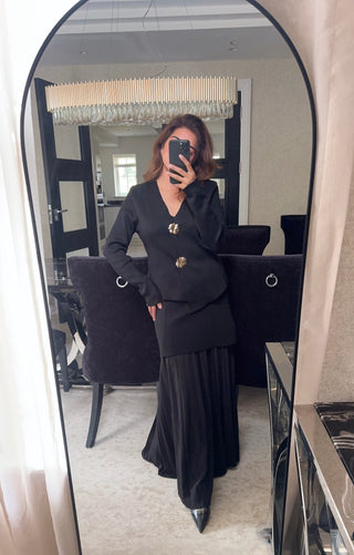 Model wearing a black double-breasted blazer with gold buttons and a pleated skirt, standing in a luxury lounge setting